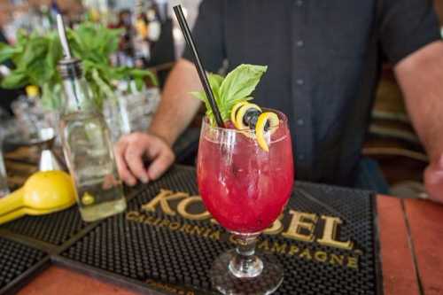 A vibrant cocktail garnished with basil and lemon twists, served in a glass on a bar counter.