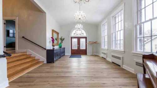 Spacious entryway with wooden stairs, large windows, chandeliers, and a decorative mirror.