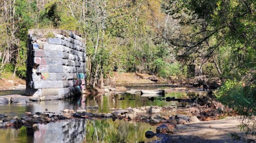 A stone bridge remnant stands by a calm stream, surrounded by trees and colorful graffiti on the stones.