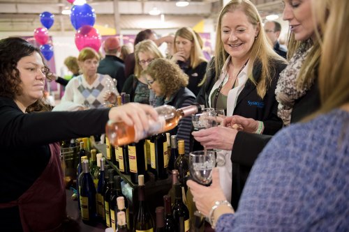 A woman pours wine for guests at a lively event with colorful balloons and a crowd in the background.