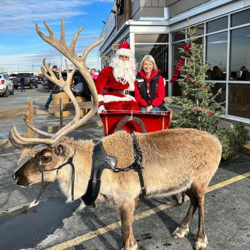 Santa Claus sits in a sleigh with a woman, while a reindeer stands nearby in front of a festive storefront.