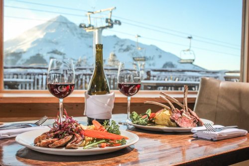 A cozy dining table with gourmet dishes, red wine, and a snowy mountain view through the window.