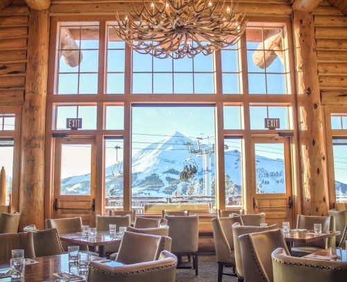Cozy restaurant interior with wooden decor, large windows showcasing a snowy mountain and ski lift outside.