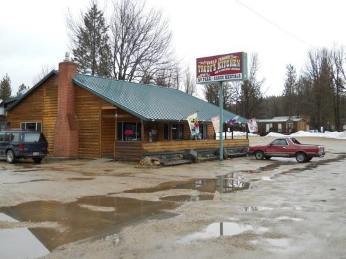 A rustic wooden building with a green roof, featuring a sign for "Trudy's Kitchen" and parked cars in a snowy area.