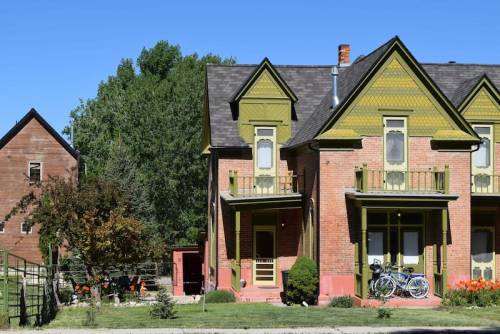 A colorful two-story brick house with green accents, surrounded by trees and a garden, featuring bicycles on the porch.