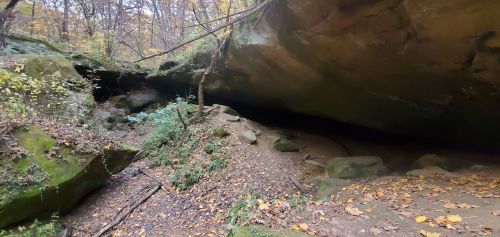 A rocky cave entrance surrounded by autumn foliage and a dirt path leading into the cave.