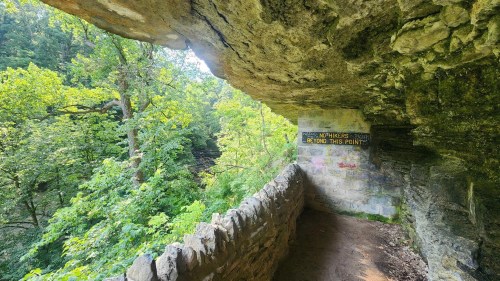 A stone overhang with a warning sign, surrounded by lush green trees and a scenic view of the forest beyond.