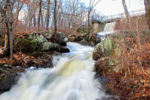 A flowing waterfall cascades over rocks, surrounded by trees and autumn leaves, under a clear blue sky.