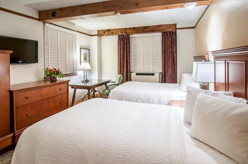 A cozy hotel room with two beds, a desk, a TV, and warm wooden accents. Natural light from a window.