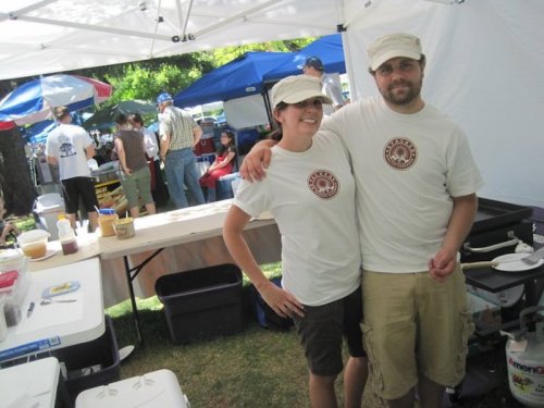 A man and woman in matching shirts and caps stand together at a food booth, with tents and people in the background.