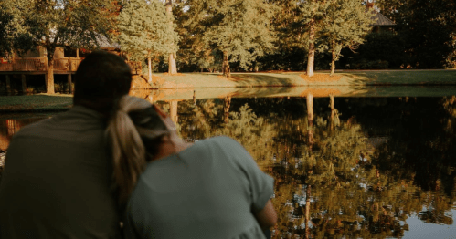 A couple sits by a serene lake, surrounded by trees, enjoying a peaceful moment together at sunset.