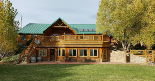 A large wooden house with a green metal roof, surrounded by trees and a grassy area, featuring a spacious deck.