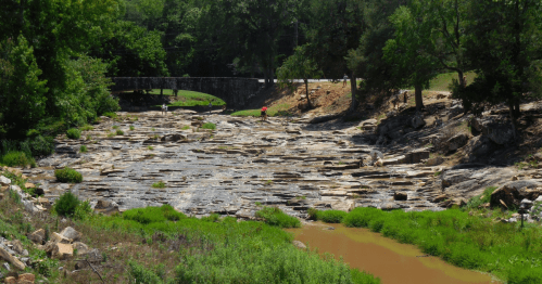 A rocky riverbed with patches of green grass, surrounded by trees and a bridge in the background.