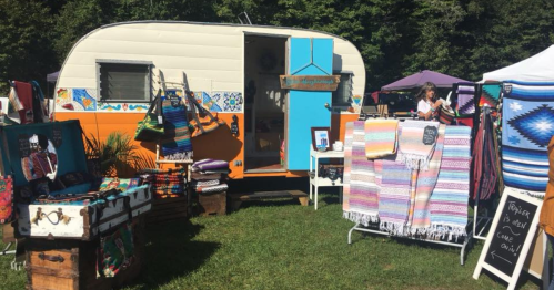 A colorful vintage trailer with handmade crafts and textiles displayed outside in a grassy area.