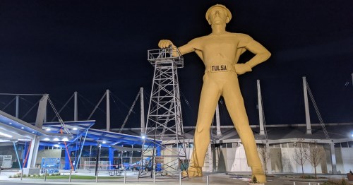 A large golden statue of a man in a hard hat stands next to a metal tower, illuminated at night in Tulsa.