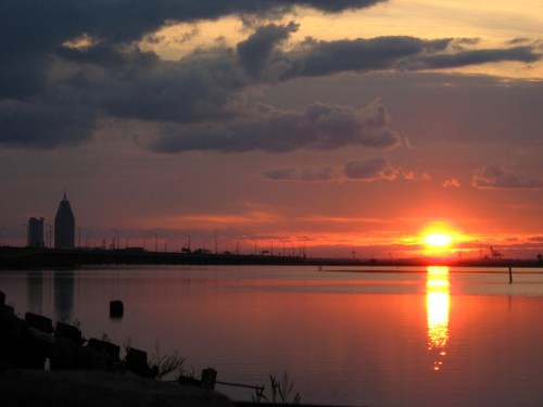 Sunset over a calm lake, reflecting vibrant orange and purple hues, with city skyline silhouettes in the background.