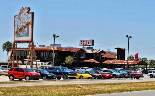 A rustic seafood restaurant with a large sign, surrounded by parked cars and palm trees under a clear blue sky.