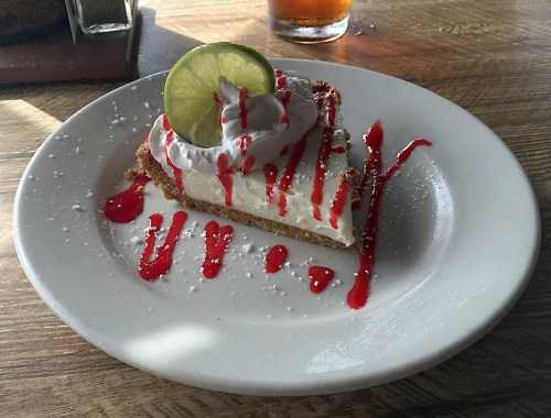 A slice of key lime pie topped with whipped cream and a lime slice, drizzled with red sauce on a white plate.