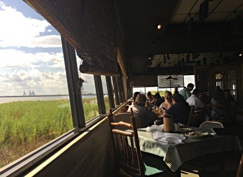 A dining area with large windows overlooking a grassy landscape and distant industrial structures, filled with patrons.