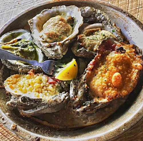 A plate of baked oysters topped with breadcrumbs, herbs, and lemon wedges, served with a fork.