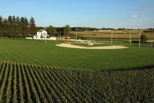 A scenic view of a baseball field surrounded by lush green grass and cornfields, with a house in the background.
