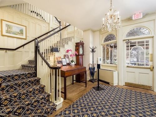 A welcoming foyer with a staircase, elegant chandelier, and decorative carpet, featuring a wooden table and coat rack.