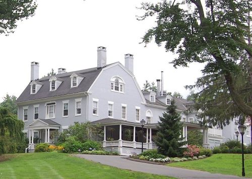 A large, elegant gray house with multiple chimneys, surrounded by lush greenery and colorful flower beds.
