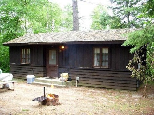 A rustic wooden cabin surrounded by trees, with a fire pit in front and a small boat nearby.