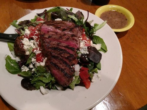A colorful salad topped with grilled steak, tomatoes, and feta cheese, served with a small bowl of dressing on the side.