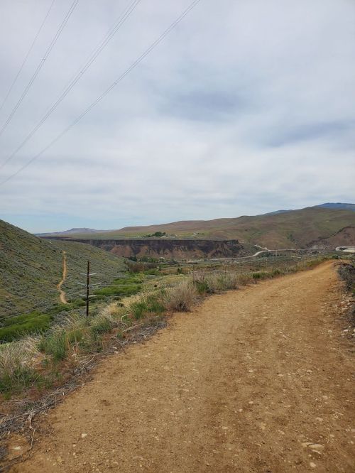 A dirt path winds through green hills under a cloudy sky, leading towards distant mountains and a valley.