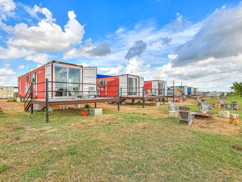 Colorful shipping container homes on raised platforms, surrounded by grassy areas and a fire pit under a blue sky.