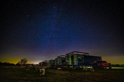 A starry night sky above a row of shipping container homes, with a faint glow on the horizon.