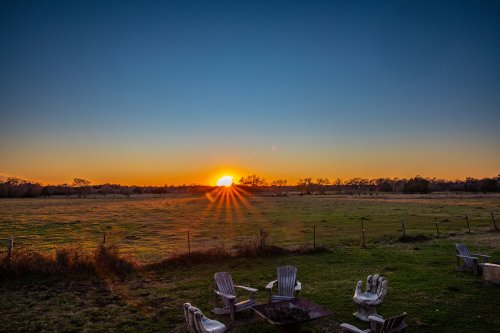 A serene sunset over a grassy field, with white chairs arranged around a fire pit in the foreground.