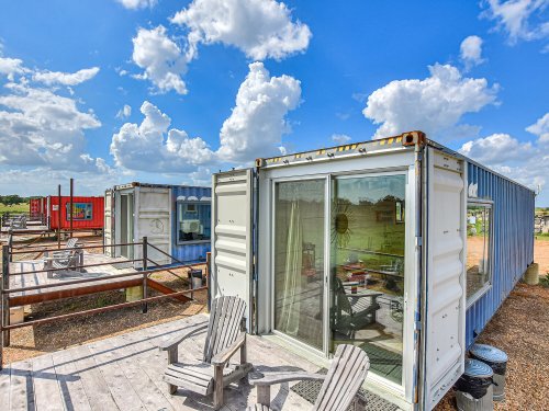 Colorful shipping container homes on a sunny day, surrounded by blue skies and fluffy clouds. Outdoor seating visible.