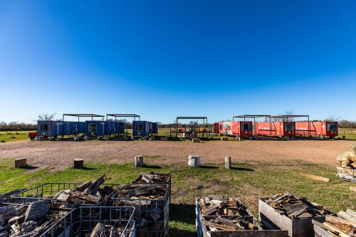 A wide view of colorful shipping containers arranged in a field, with firewood stacked in bins in the foreground.