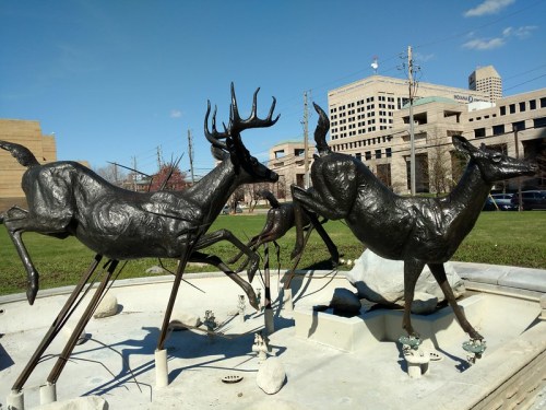 Sculpture of running deer in a park, with buildings and blue sky in the background.