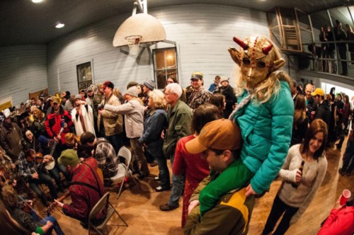 A lively gathering in a hall with people dancing, playing instruments, and a child in a mask on someone's shoulders.