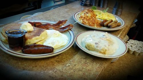 A hearty breakfast spread featuring pancakes, bacon, eggs, sausage, hash browns, and a cinnamon roll with icing.
