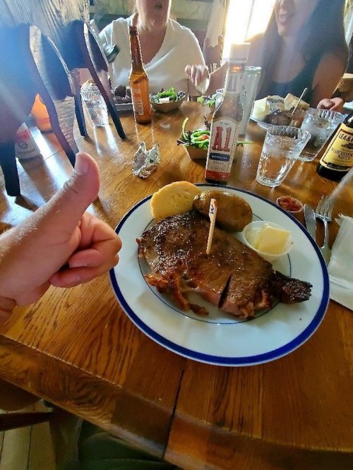 A hand gives a thumbs up next to a plate of steak, potatoes, and condiments on a wooden table with people in the background.