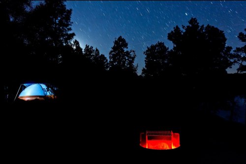 A serene night scene with a glowing campfire and a tent under a starry sky, surrounded by trees.
