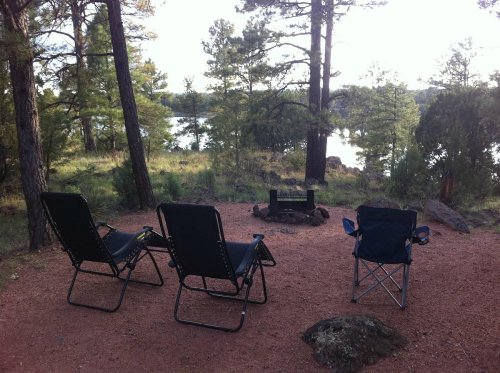Three camping chairs are set up near a fire pit, surrounded by trees and overlooking a serene lake.