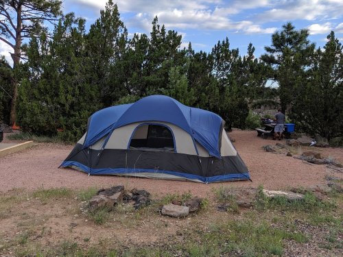 A blue and gray tent set up in a gravel campsite surrounded by trees and a distant figure at a picnic table.