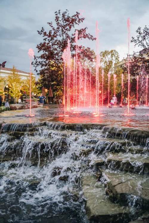 A vibrant fountain with colorful jets of water, surrounded by trees and people enjoying the evening atmosphere.