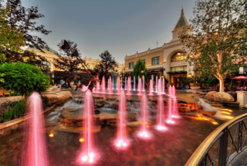 Colorful fountains with pink lights in a landscaped area, surrounded by trees and elegant buildings at dusk.