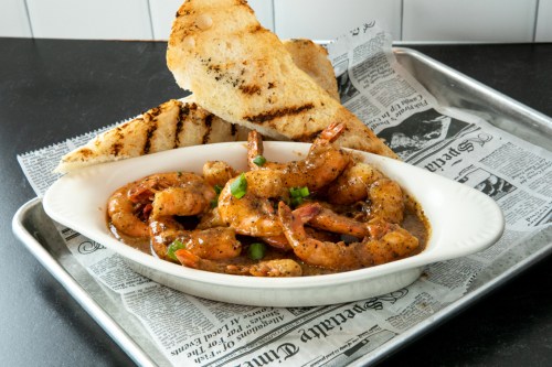 A plate of shrimp in a spicy sauce, garnished with green onions, served with grilled bread on a tray lined with newspaper.