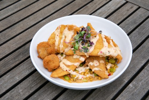 A white bowl filled with fried fish, corn, and green peas, topped with a creamy sauce and microgreens, served on a wooden table.