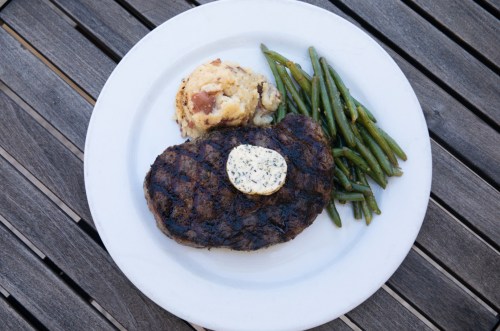 A grilled steak topped with herb butter, served with mashed potatoes and green beans on a white plate.