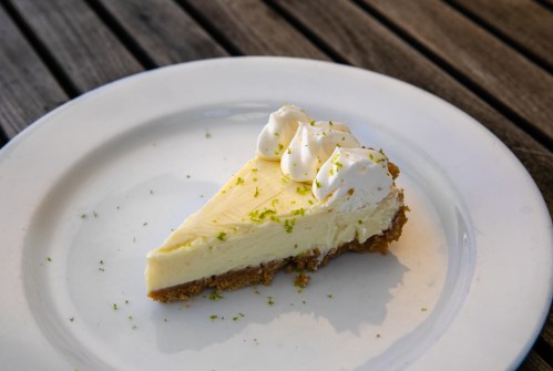 A slice of key lime pie topped with whipped cream and lime zest on a white plate, resting on a wooden table.