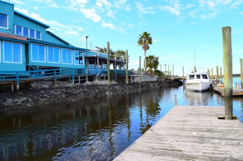A calm marina scene with a boat docked beside a wooden pier, surrounded by palm trees and a blue building.