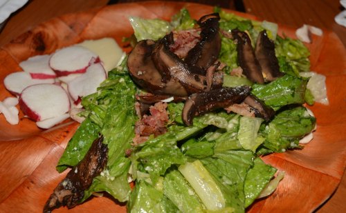 A wooden plate with a fresh salad of romaine lettuce, mushrooms, bacon, and sliced radishes on the side.